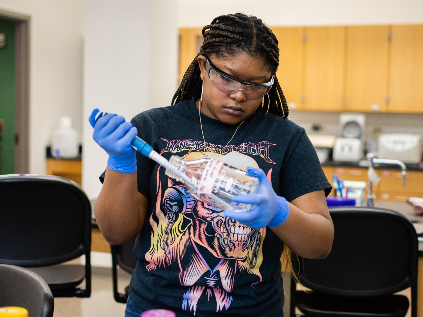 A student, wearing lab goggles and gloves, uses a pipette in a chemistry lab.